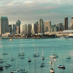 A picturesque view of the San Diego skyline with sailboats anchored in the harbor under a cloudy sky.