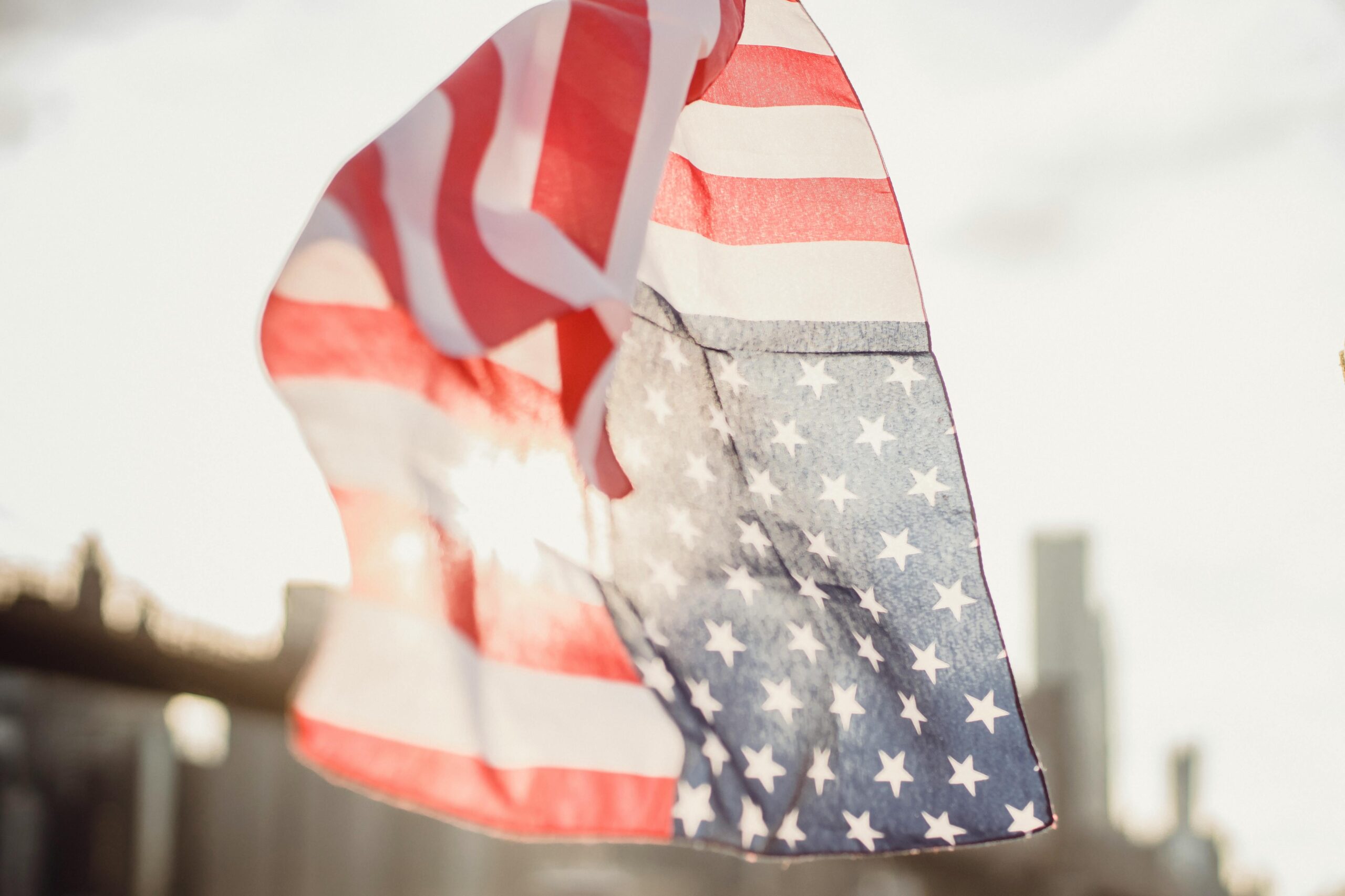 Close-up of an American flag waving against a sunny blurred city background.