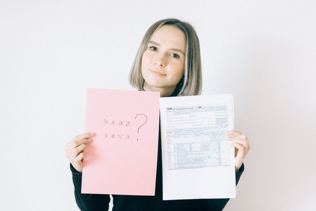A woman in a black shirt holds tax forms and a 'Need Help?' sign, indoors.